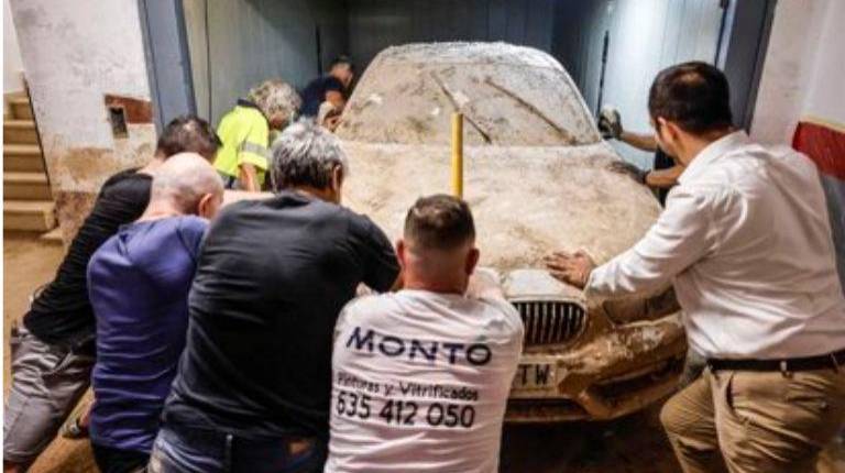 <i>Varias personas sacan uno de los coches destrozados por la dana de octubre de 2024 de un garaje en Aldaia. / Rober Solsona / Europa Press.</i>